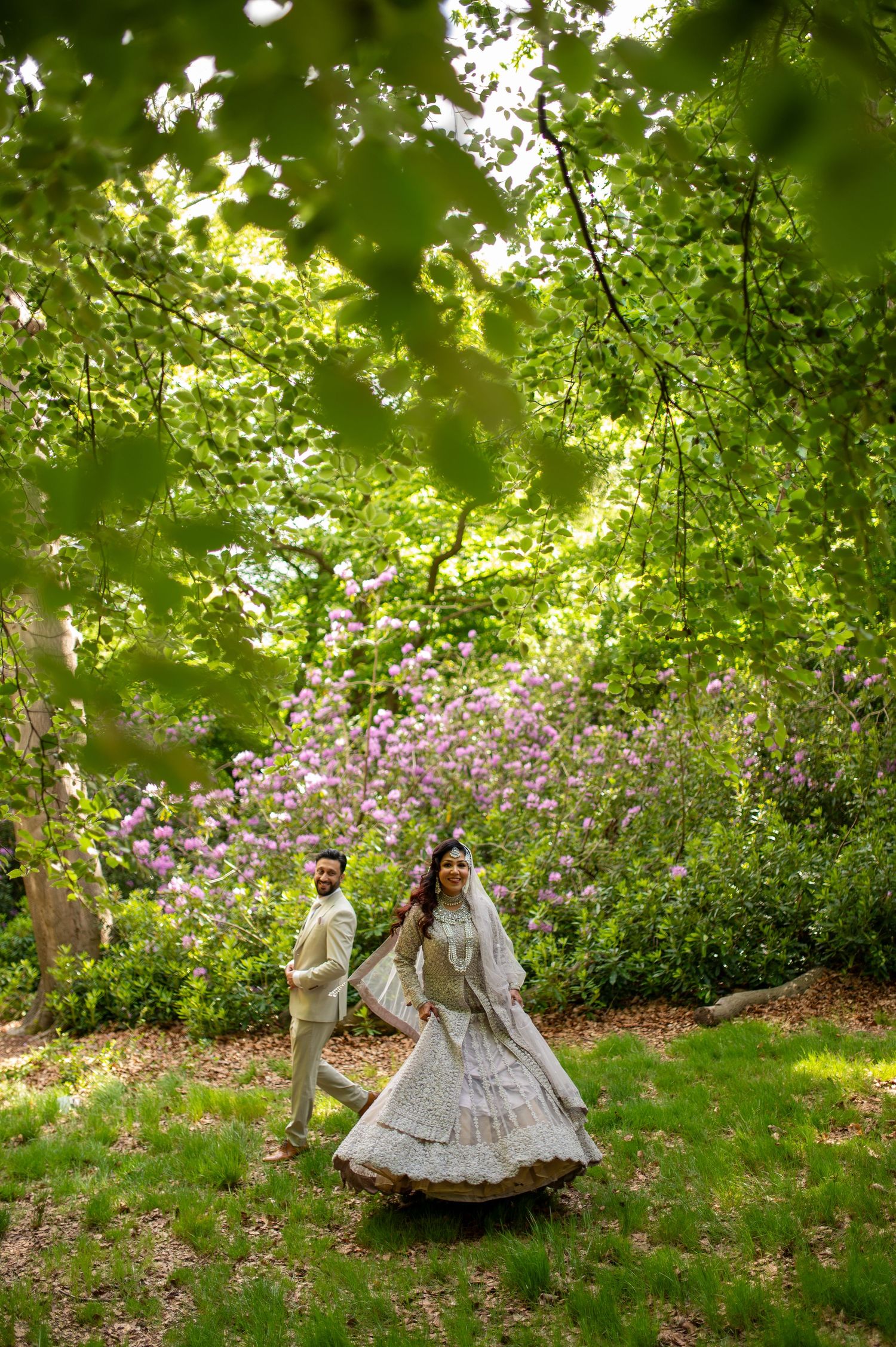 South Asian bride in ivory lehenga twirls with groom near blooming flowers in lush garden in london.