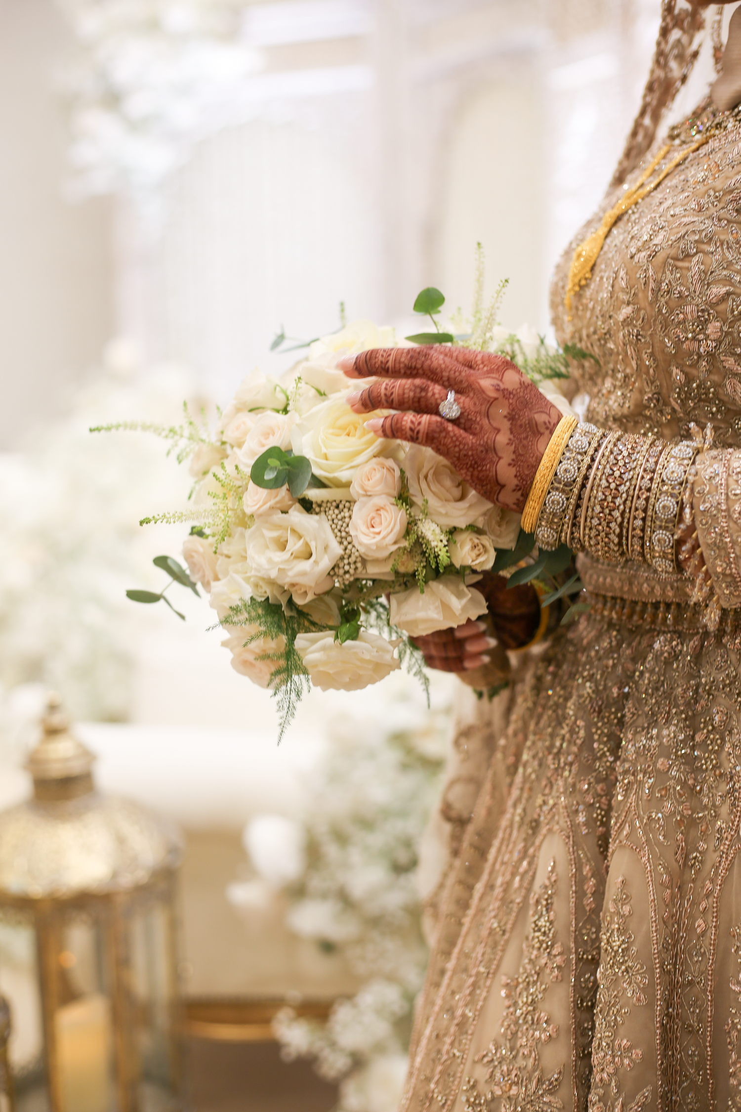 Indian bride holding white rose bouquet with henna hands, gold bangles, and embroidered lehenga in london.