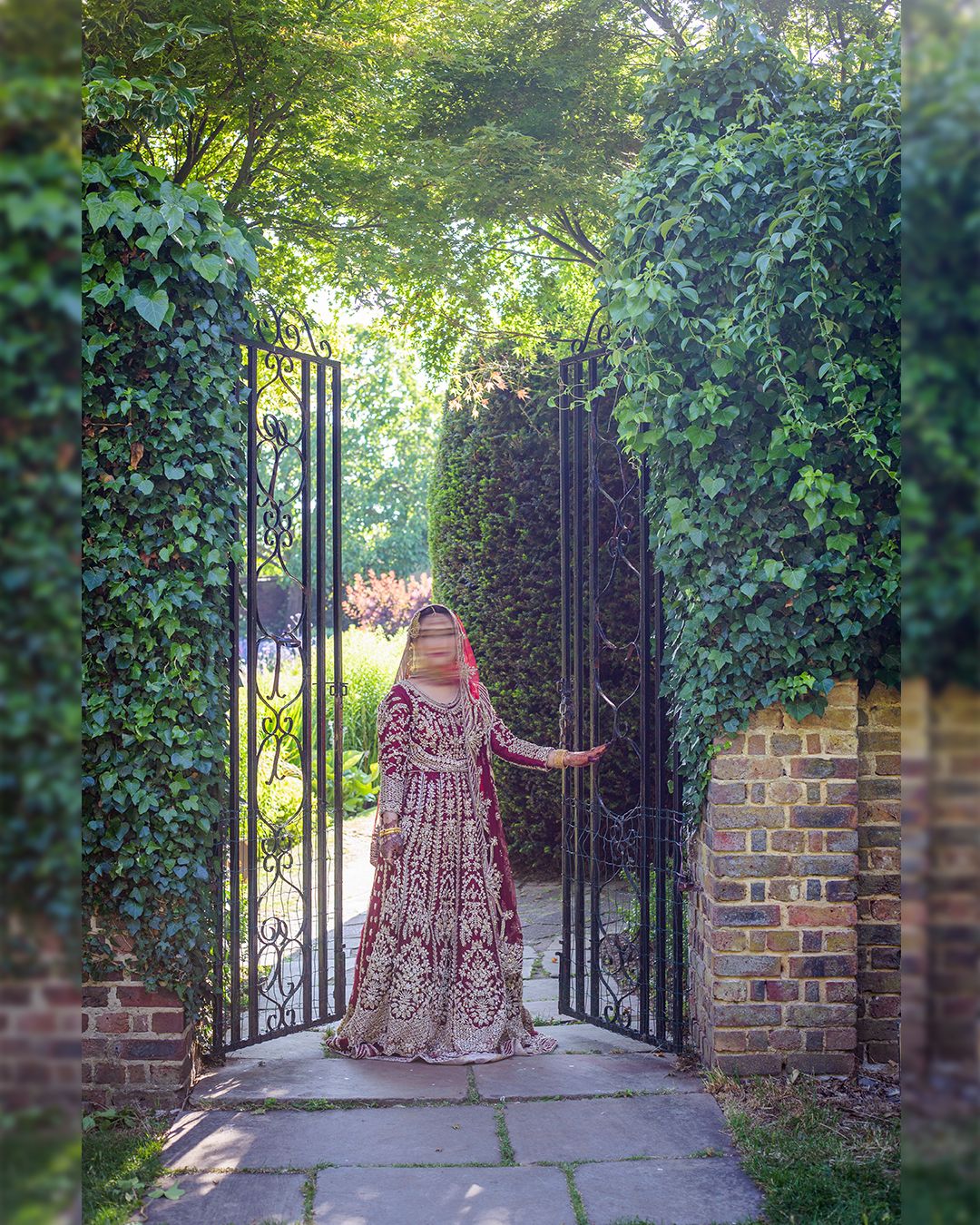 Pakistani Bride in red and gold lehenga standing at ornate iron garden gate surrounded by lush green ivy and trees in London.
