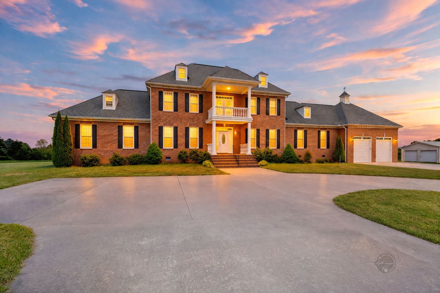 Grand brick colonial mansion with balcony and circular driveway photographed at sunset with colorful sky.