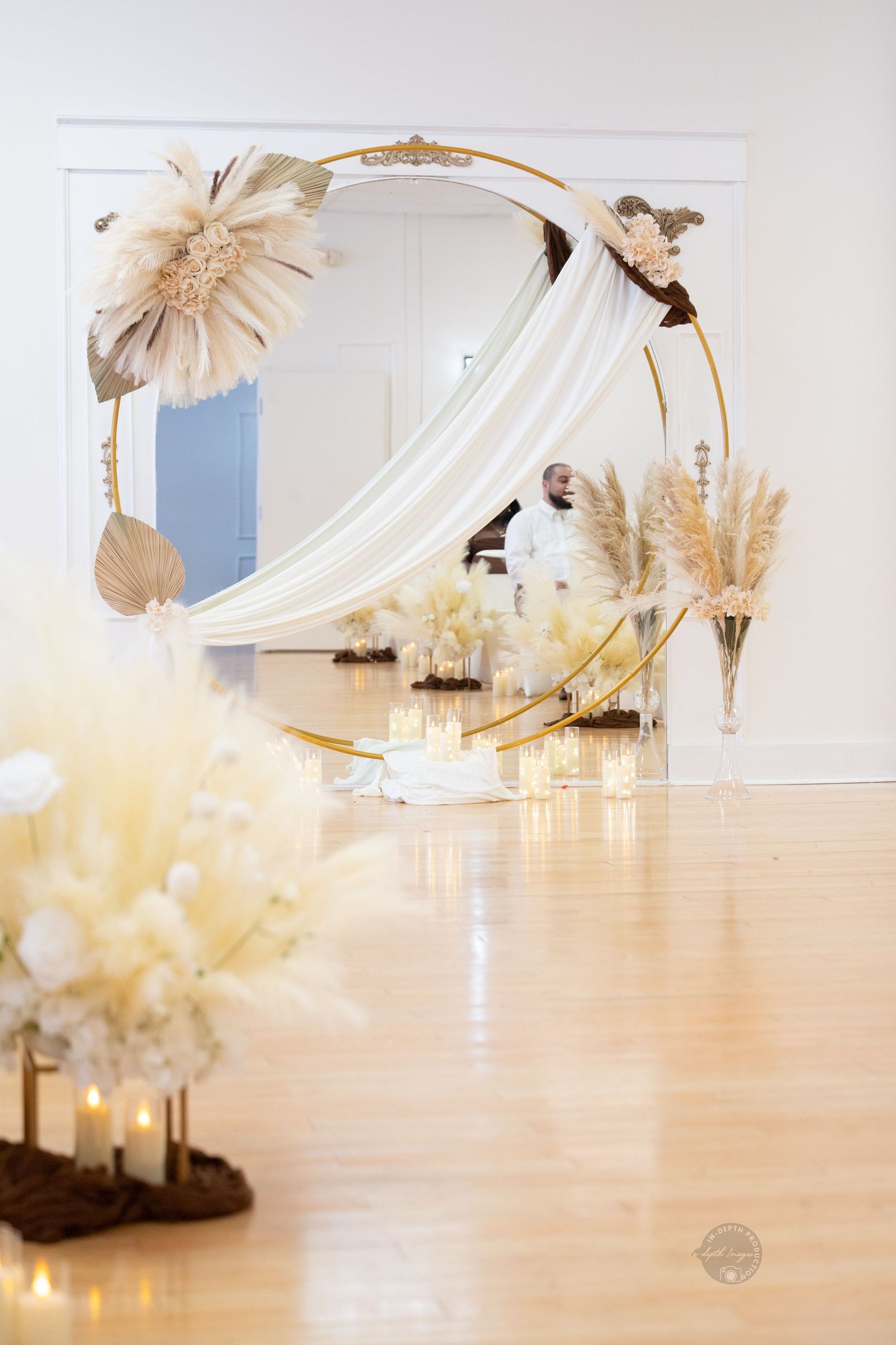 Elegant gold circular wedding arch with white draping, dried pampas grass, and candles on a hardwood floor.
