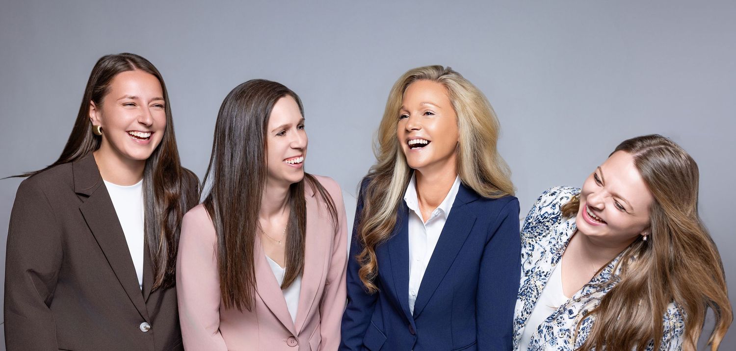 Four professional women in business attire laughing and smiling together against a gray background.