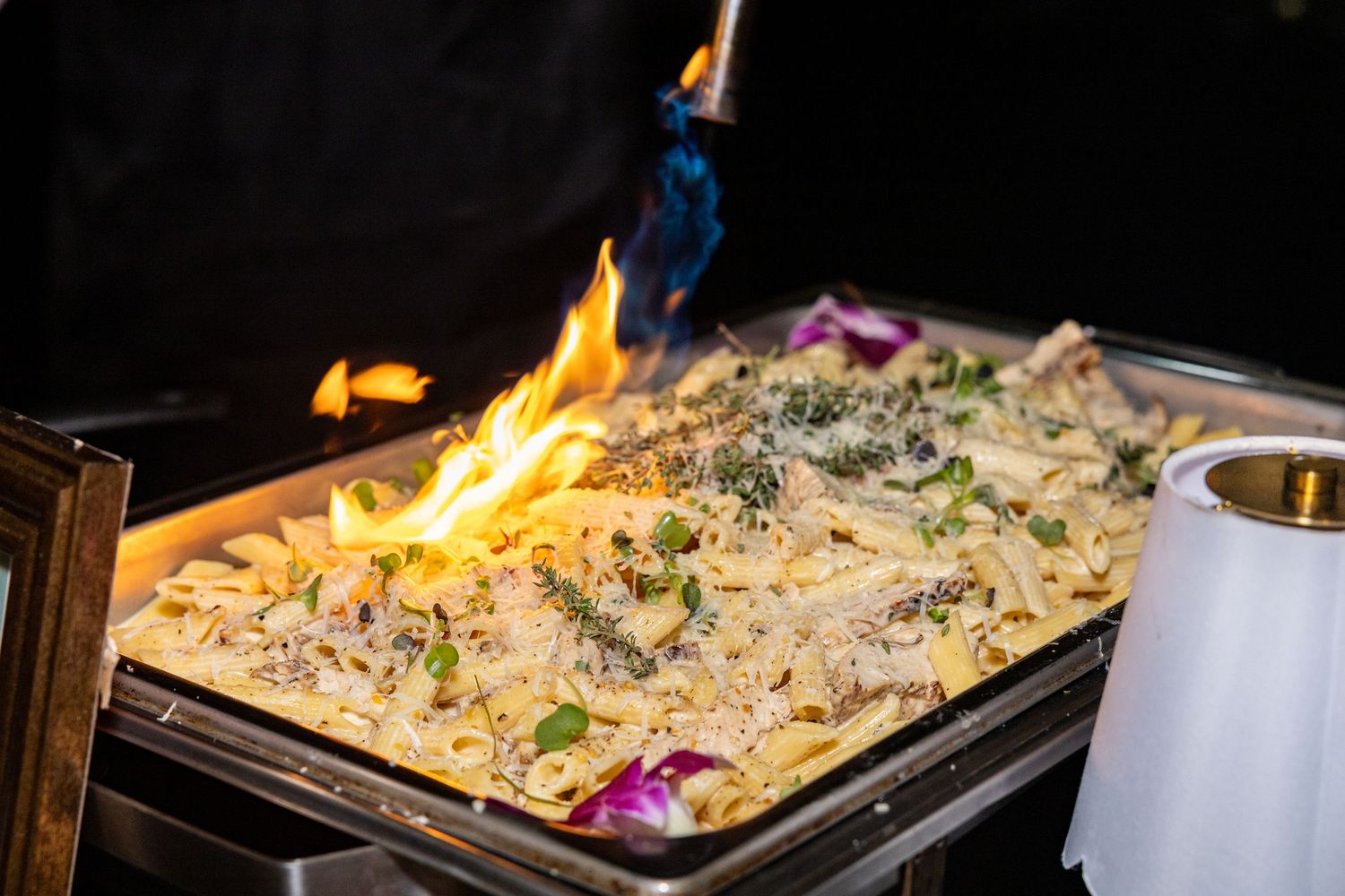 Chef flambeing creamy pasta dish in a chafing tray with blue torch flame at catered event.