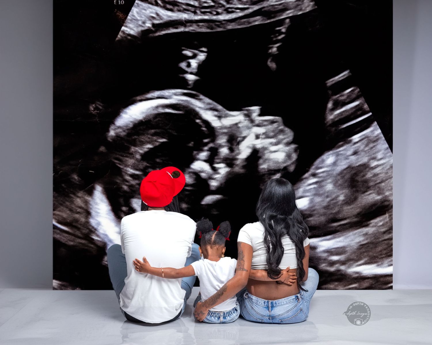 Family of three sitting together viewing a large ultrasound photo backdrop, pregnancy announcement moment.