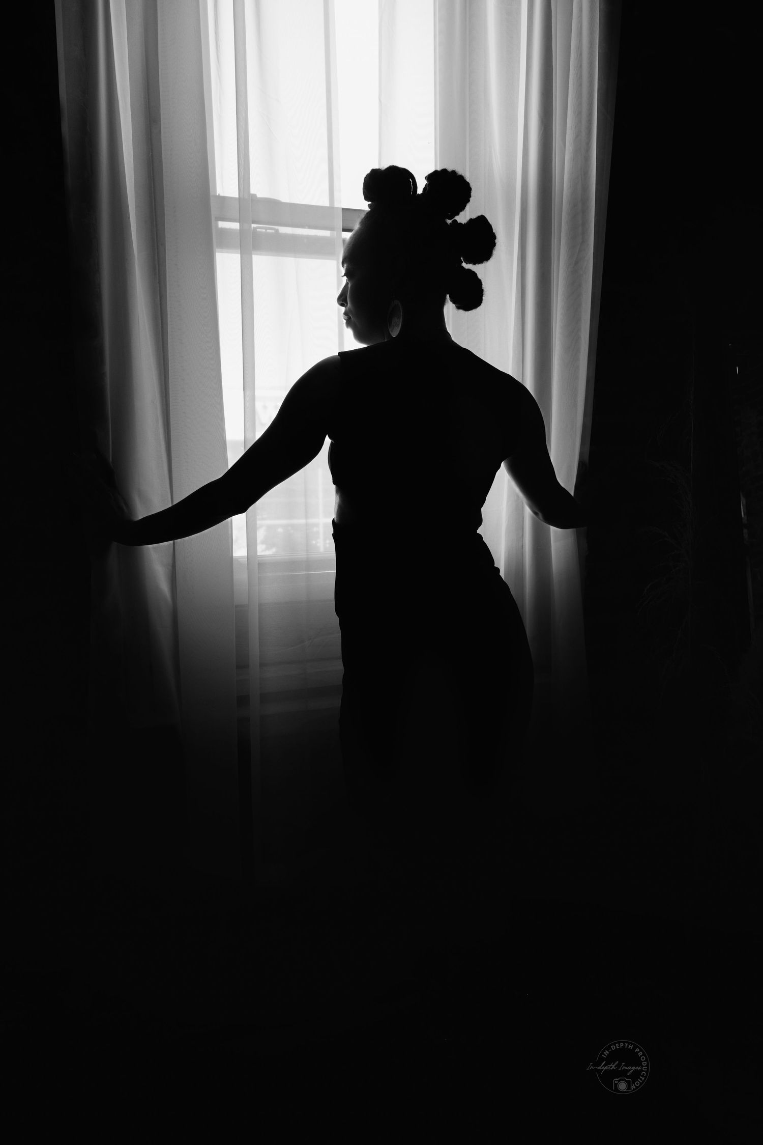 Black and white silhouette of a woman with an elegant updo hairstyle standing in front of a bright window with curtains.
