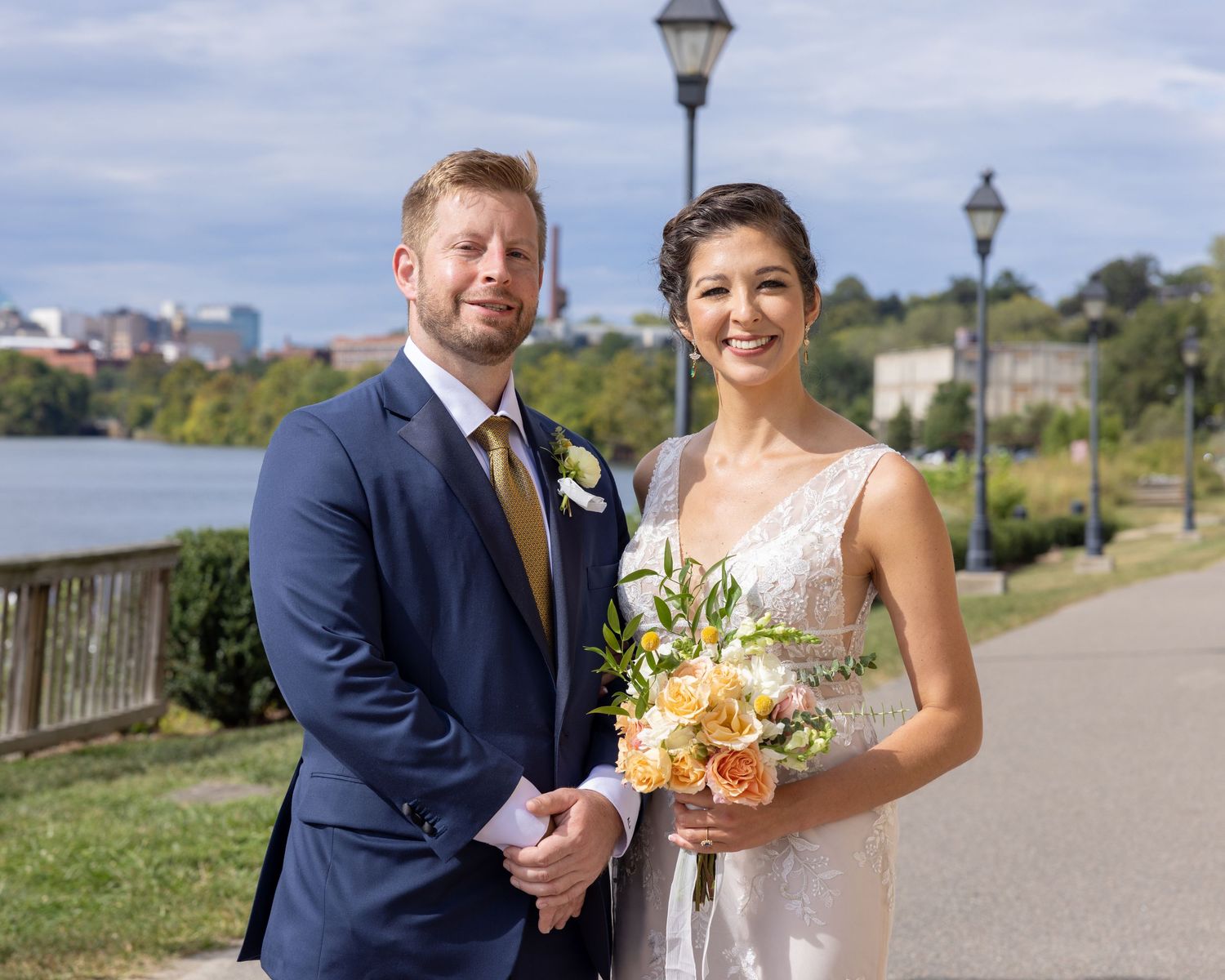 Smiling bride and groom pose on a riverside walkway, bride holding peach bouquet, groom in navy suit.