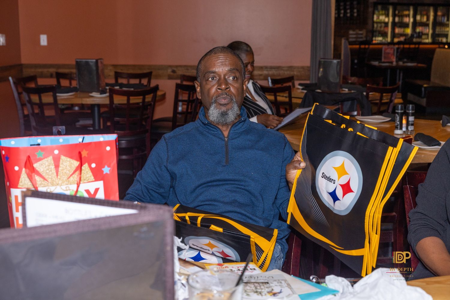 Smiling man with a gray beard sits at a restaurant table beside a black and gold Pittsburgh Steelers bag and a red gift bag.
