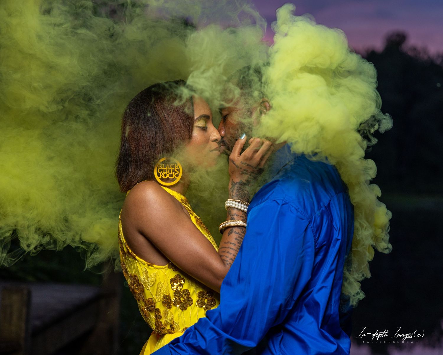 Couple kissing romantically surrounded by vibrant yellow-green smoke bombs outdoors at dusk, wearing blue and yellow outfits.