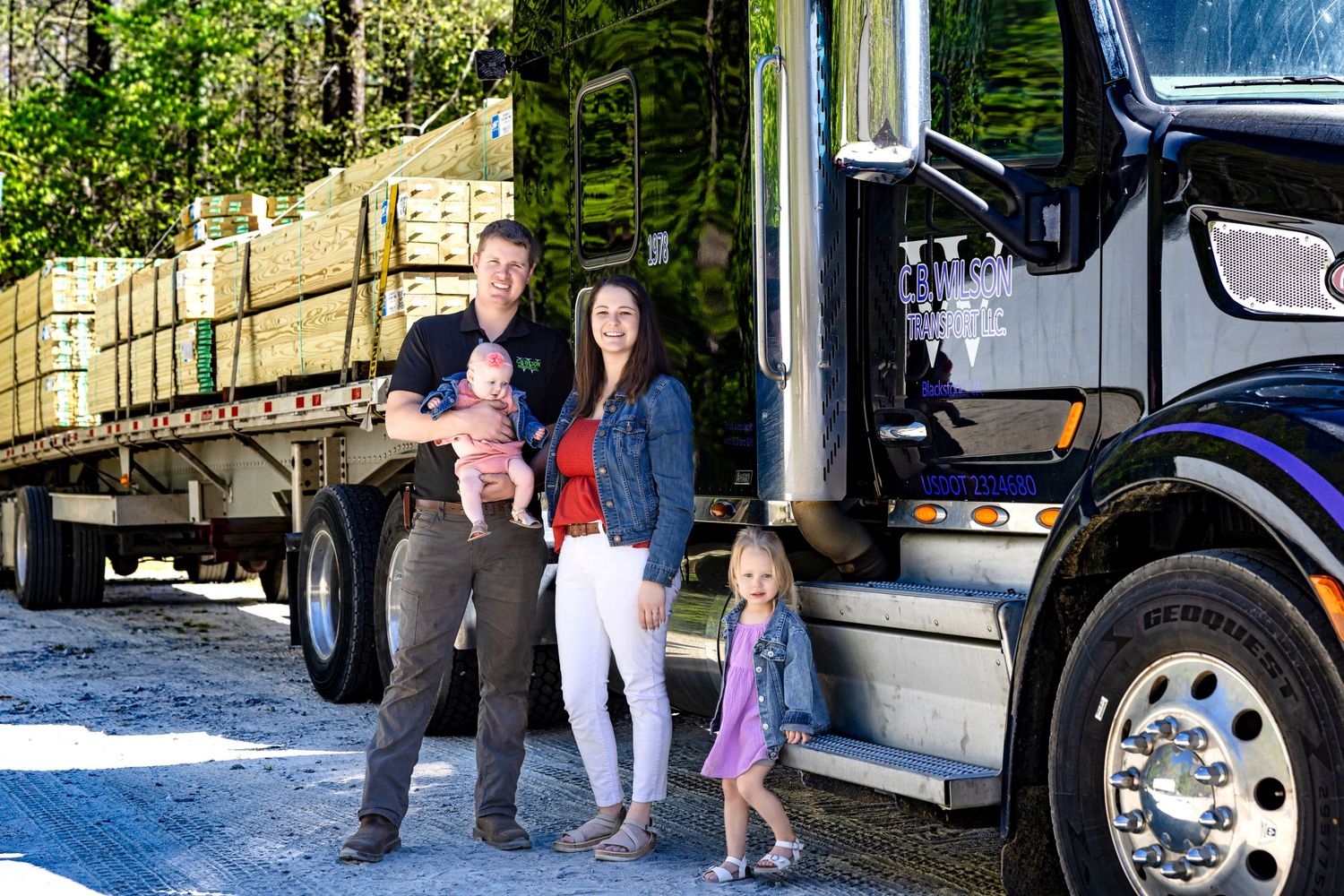 A smiling family with two young children stands in front of a large semi-truck on a gravel lot.