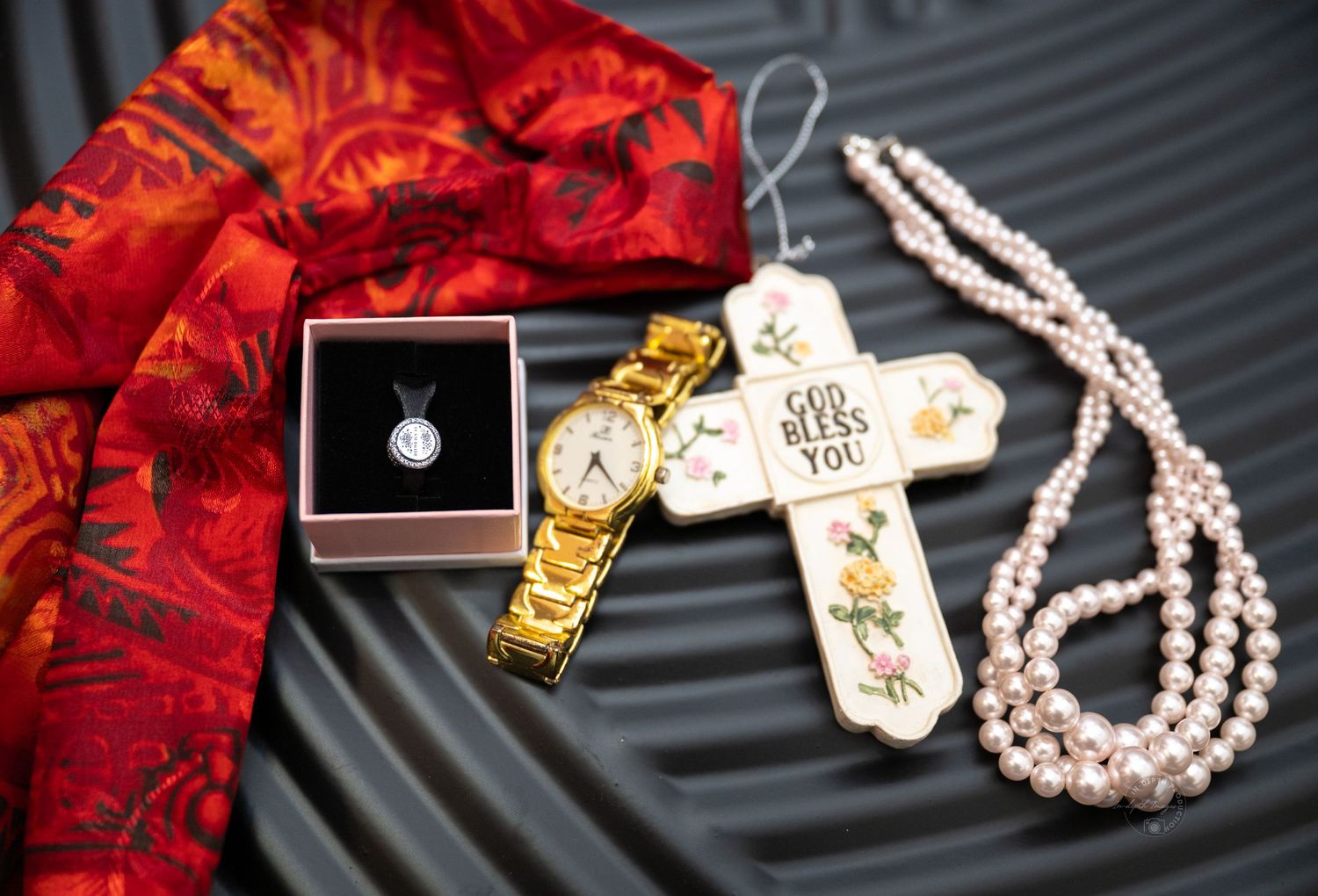 Gold watch, pearl necklace, cross ornament, and pendant necklace in box arranged on red fabric.