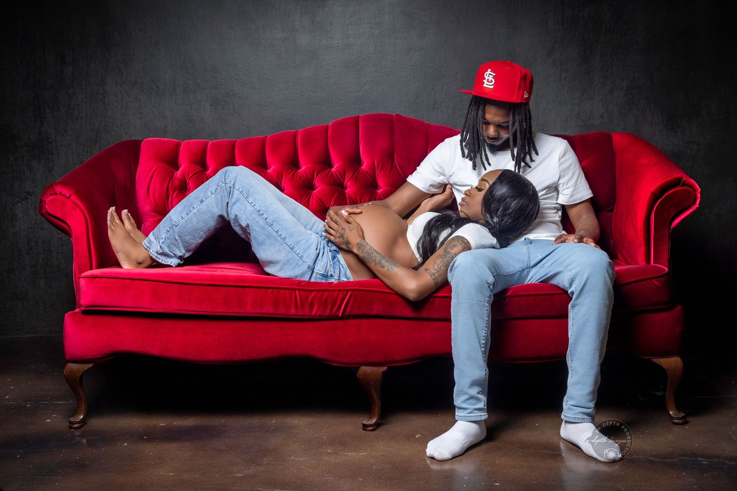 Couple relaxing on a red velvet sofa, man in white shirt and red cap, woman lying across his lap.