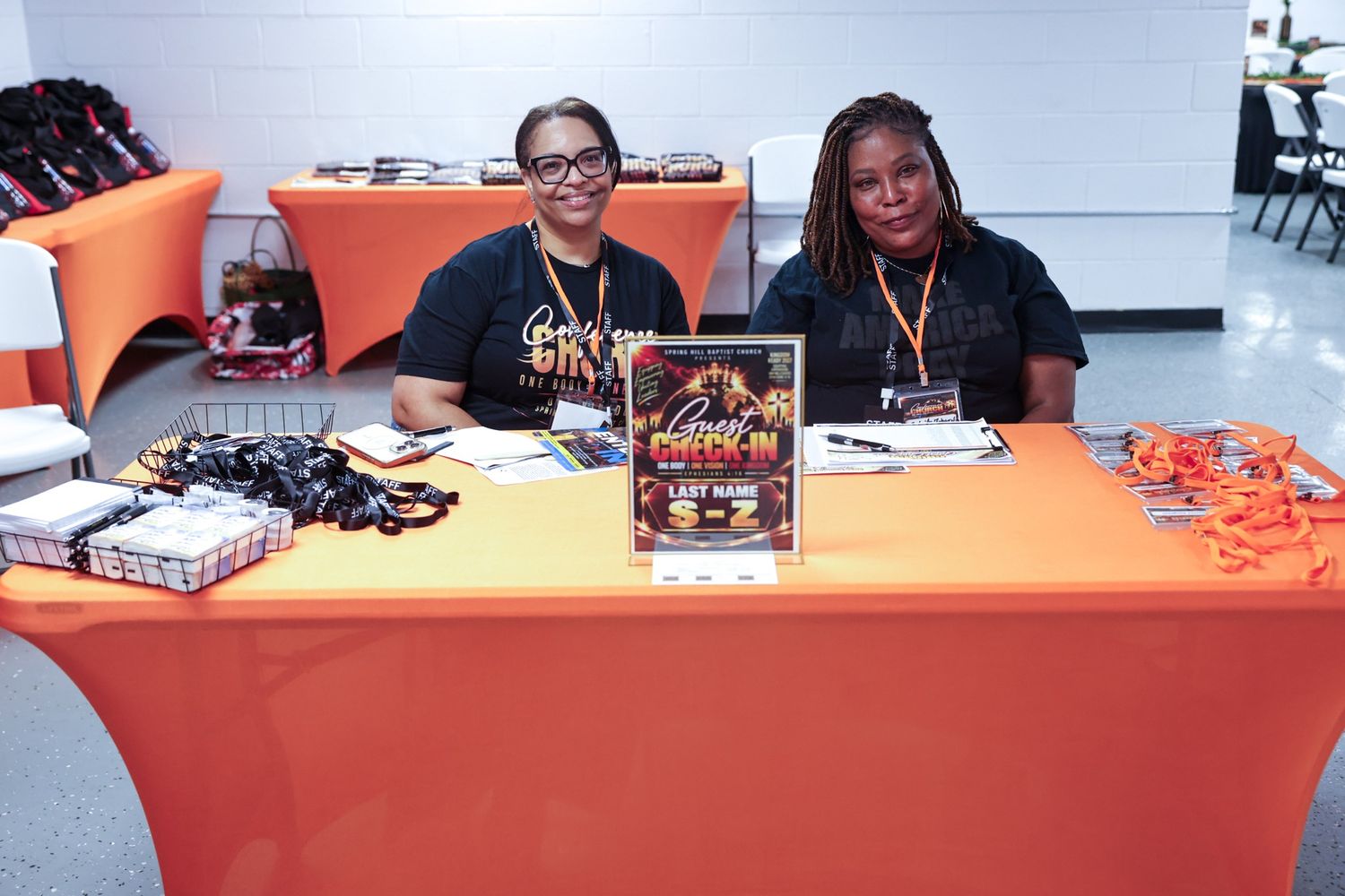 Two smiling women in black shirts sit behind an orange-draped registration table with a welcome flyer, name badges, and event supplies.