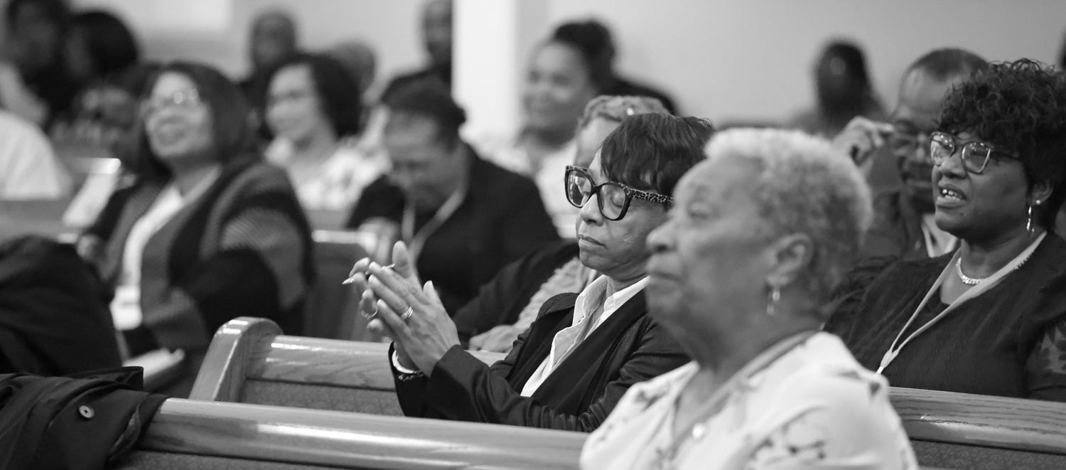 Black and white photo of congregation members seated in church pews, with a man in glasses gesturing as he listens during a service.