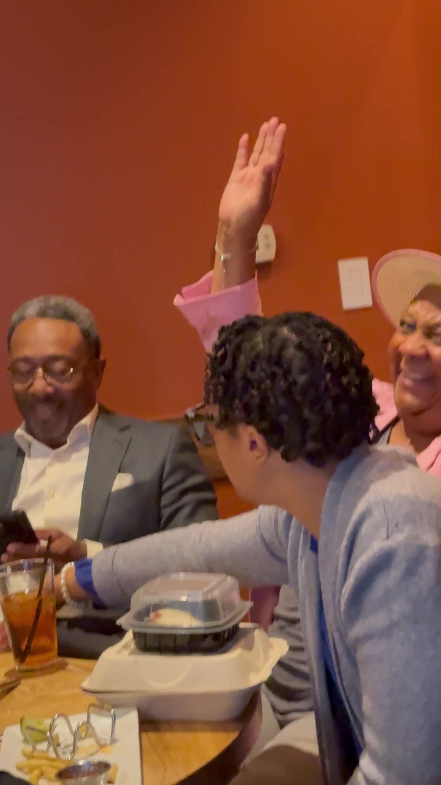 Smiling diners at a restaurant table laugh together as one person raises a hand, with drinks and takeout containers on the table.