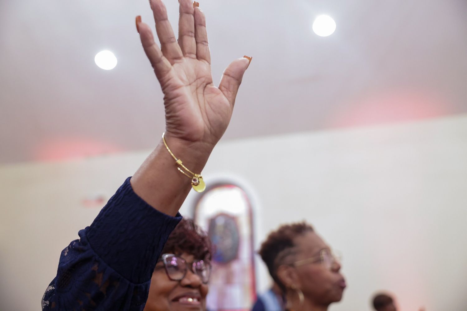 Woman with glasses raises her hand in worship during a church service while another woman stands behind her in a softly lit sanctuary.