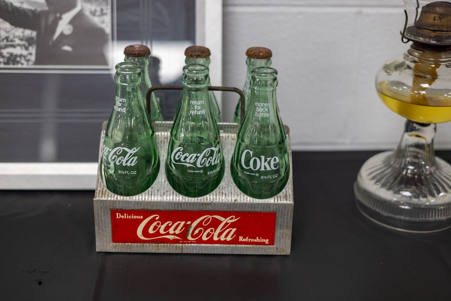 Vintage Coca-Cola glass bottles in a retro metal carrier displayed next to an antique oil lamp.