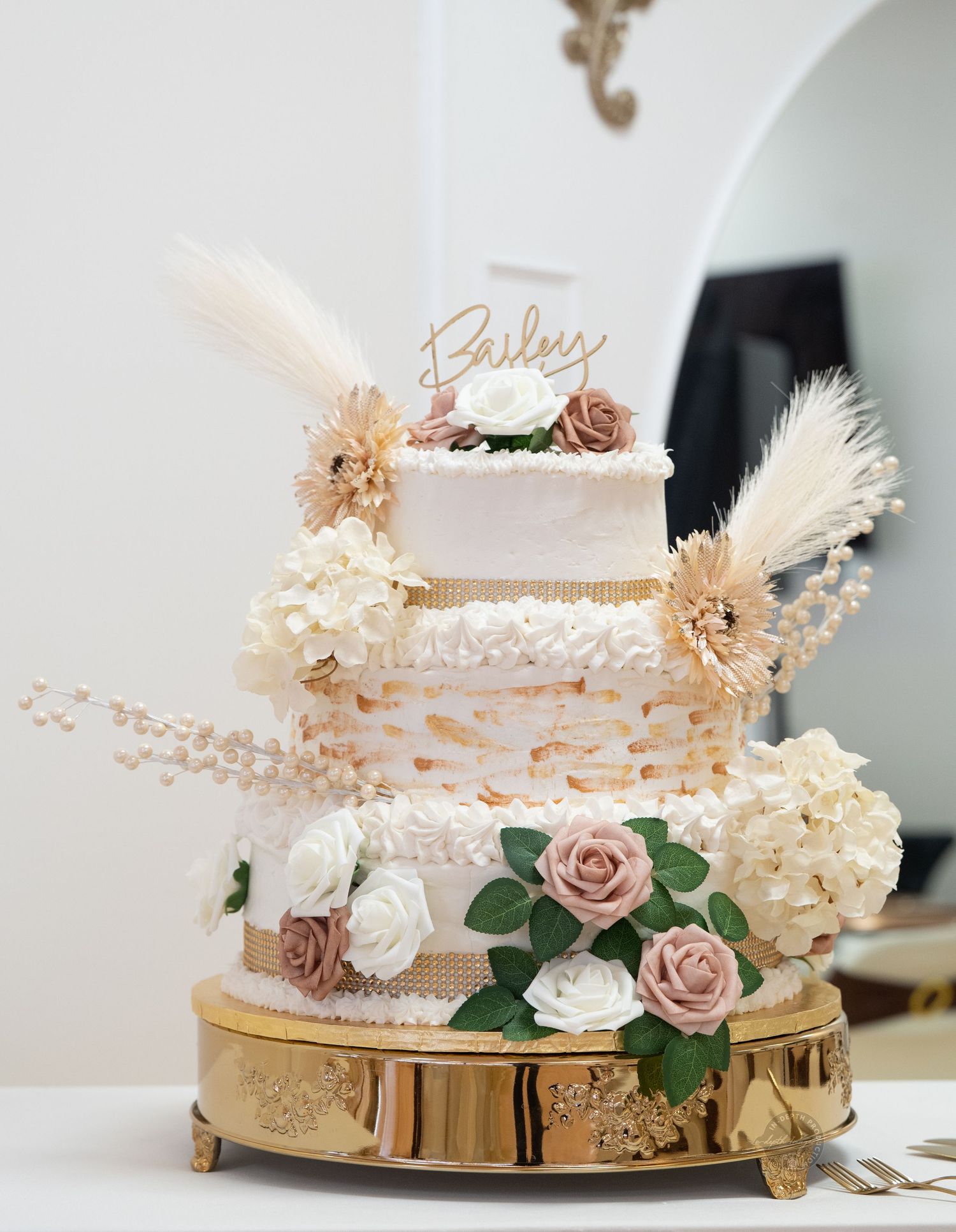 Elegant three-tier wedding cake with blush roses, white hydrangeas, pampas grass, and gold accents on a golden stand.