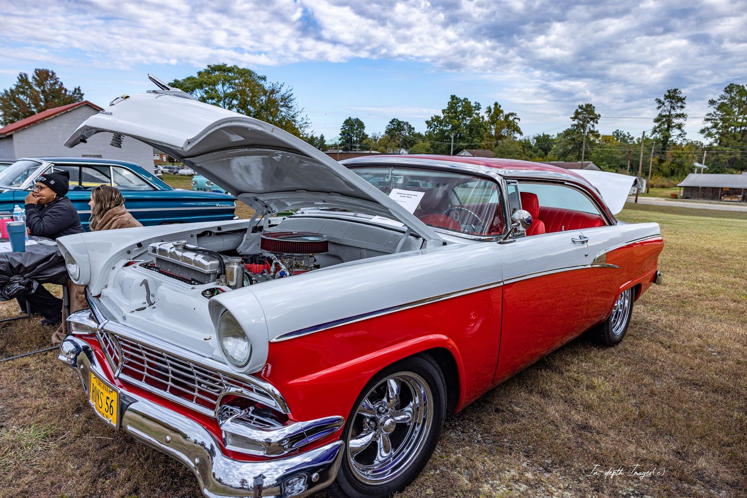 A restored red and white 1956 Ford Fairlane classic car with hood open at an outdoor car show.