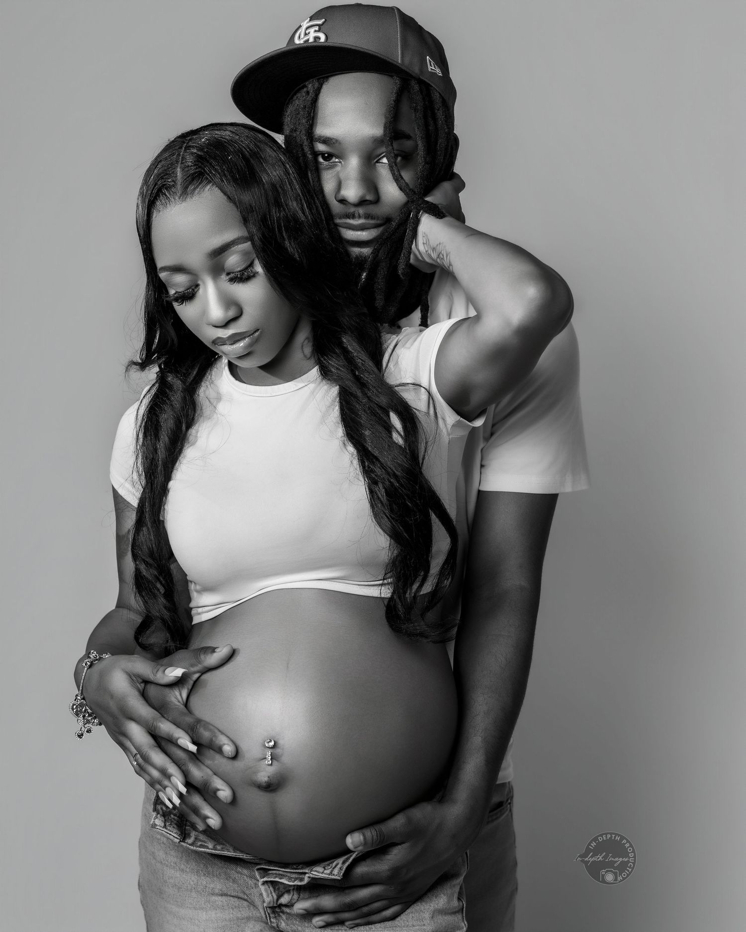 Black and white maternity photo of a pregnant couple, man embracing woman from behind, both cradling her baby bump.