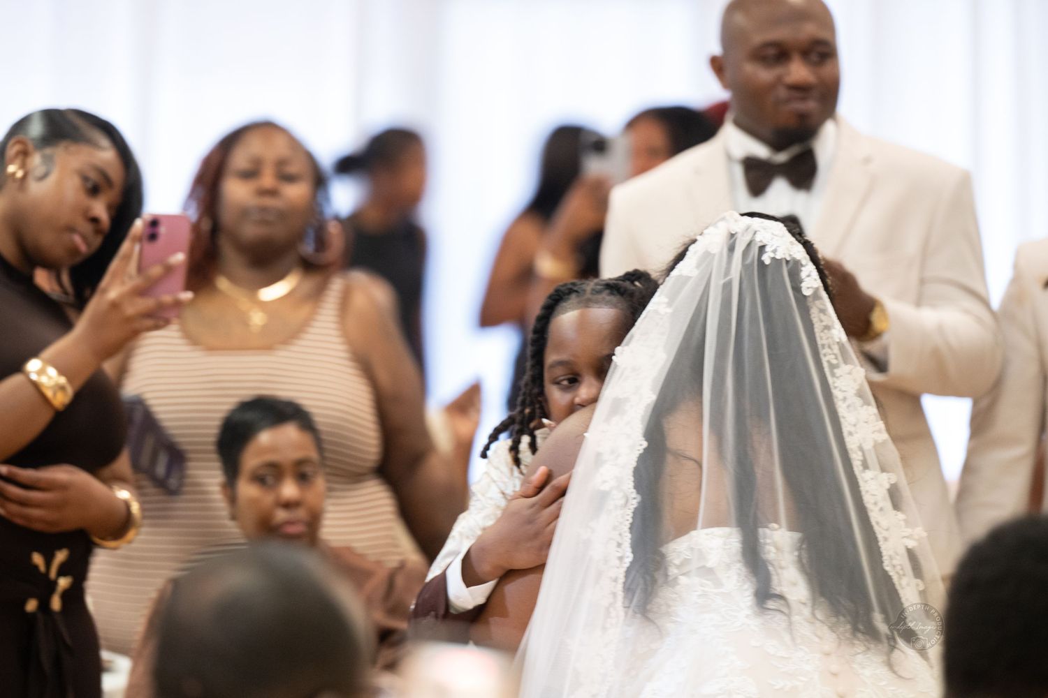 A young girl peeks out from behind a bride's veil during a wedding ceremony surrounded by guests.