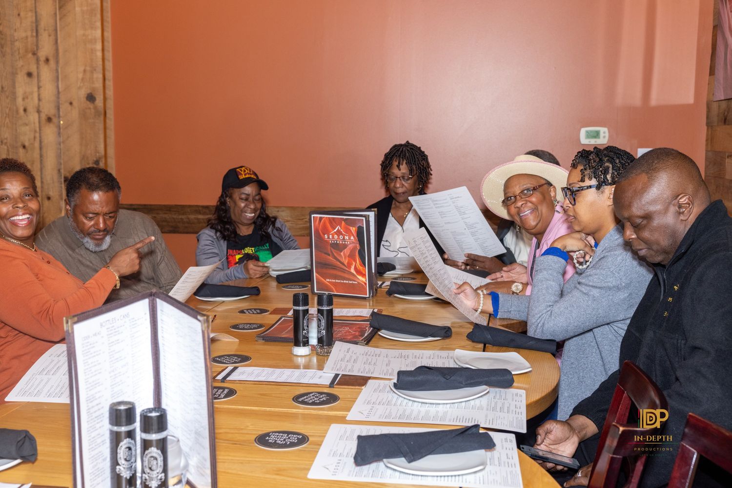 Group of smiling friends gathered around a wooden restaurant table with menus, napkins and place settings, sharing a meal together.