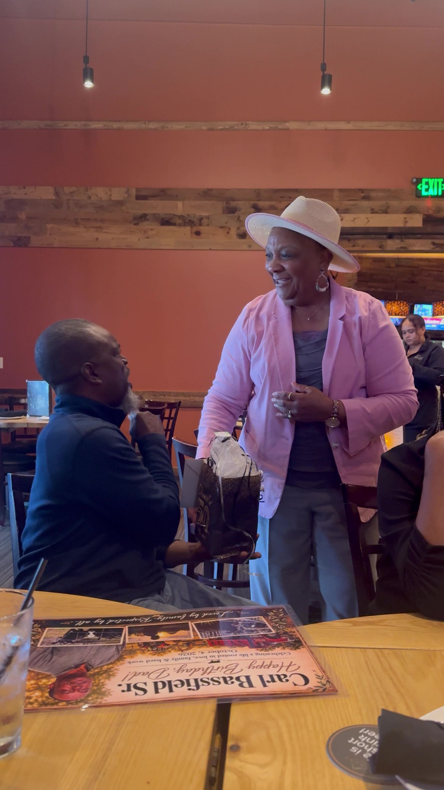 Woman in a white hat and pink blazer smiles while greeting a seated man at a restaurant table with a birthday placemat.