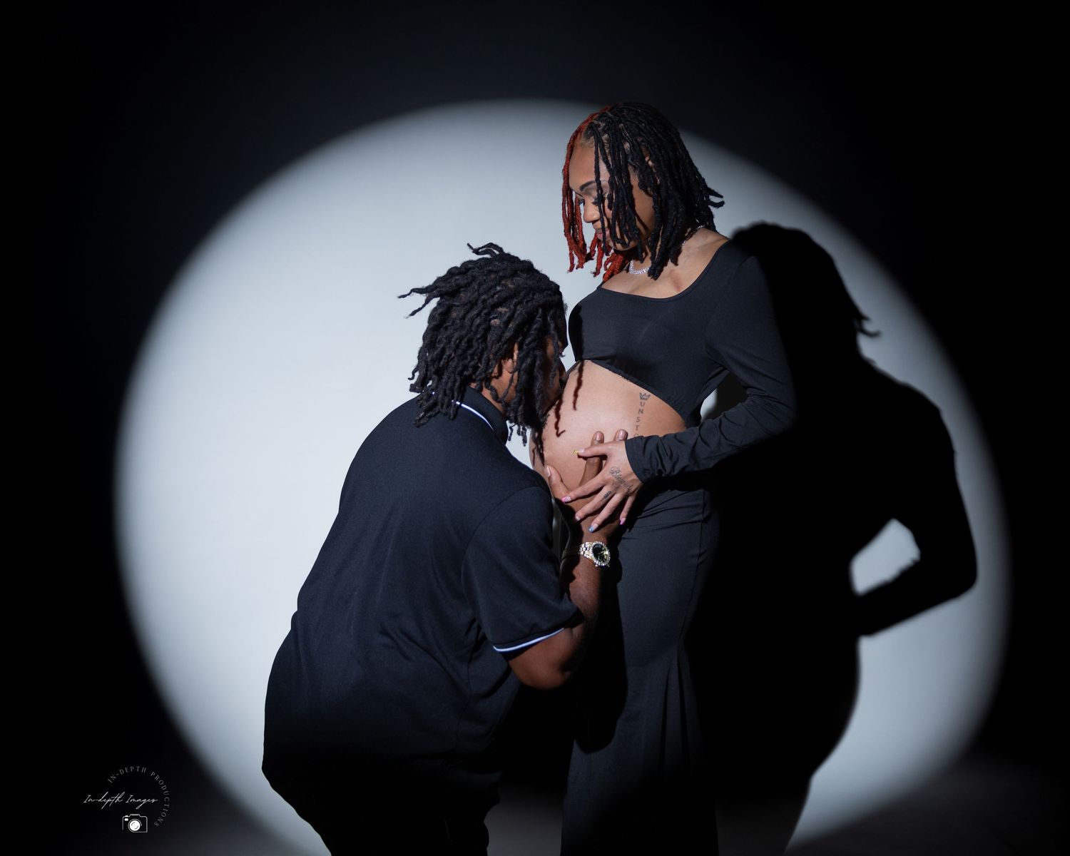 Dramatic maternity photo of couple in black outfits posed under a spotlight.