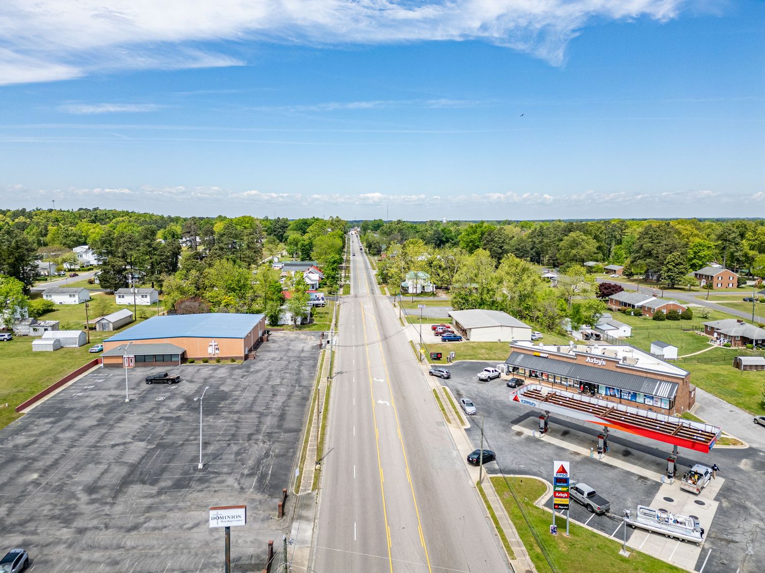 An aerial view of a straight road lined with trees, businesses, and a gas station under a blue sky.