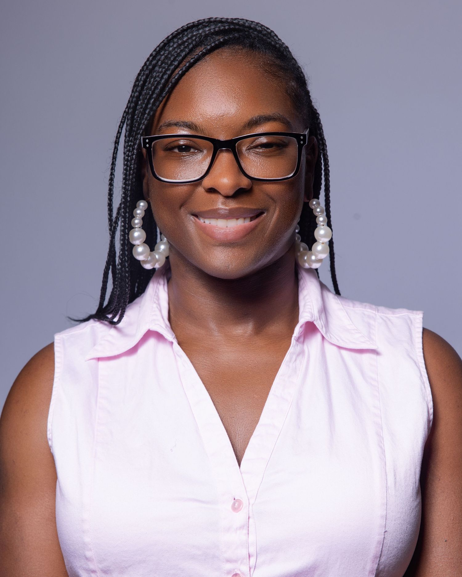 Young woman with braids and glasses wearing a pink sleeveless shirt and pearl earrings, smiling confidently.
