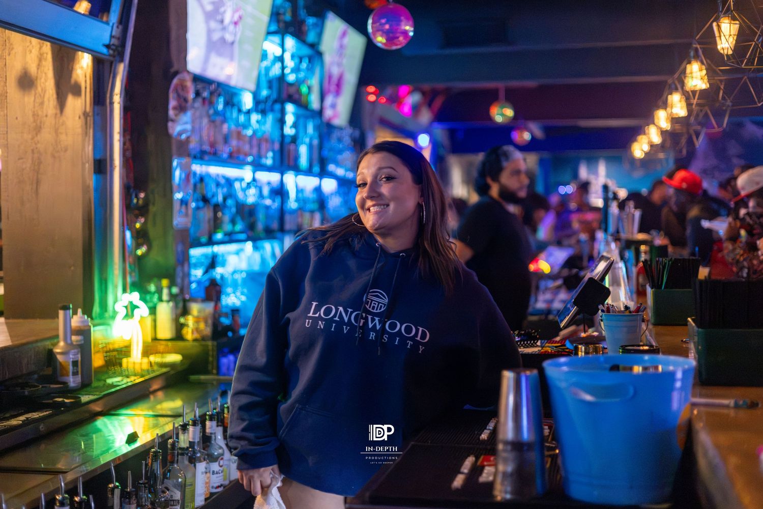 Smiling bartender in Longwood University hoodie stands behind a colorful bar with neon lights.