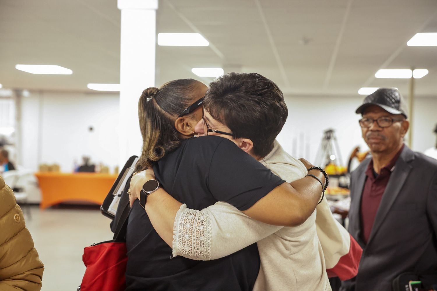 Two people share a warm embrace in a bright indoor event space while a man in a cap and glasses looks on nearby.