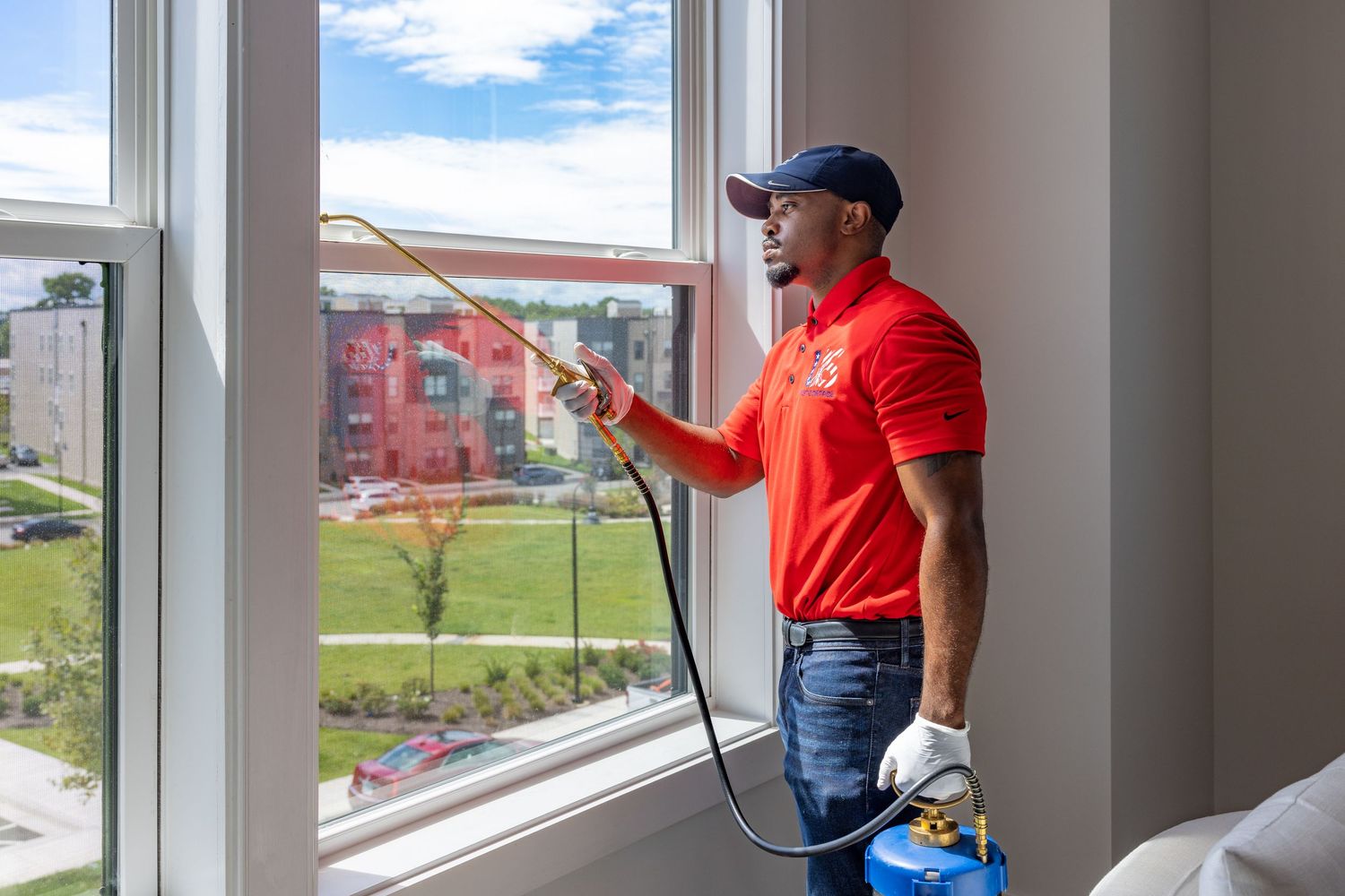 Pest control technician in red uniform spraying window interior with professional equipment in a modern apartment.