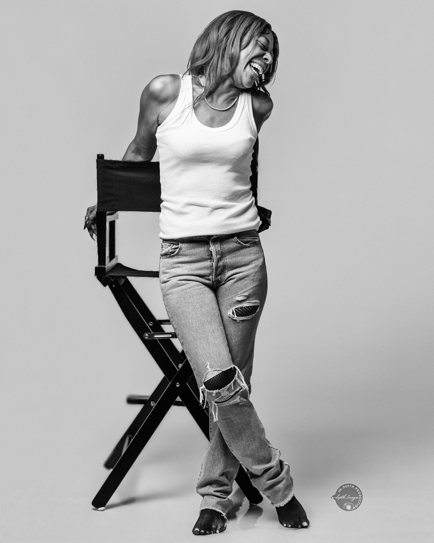 Black and white photo of a woman in ripped jeans and tank top laughing beside a director's chair.