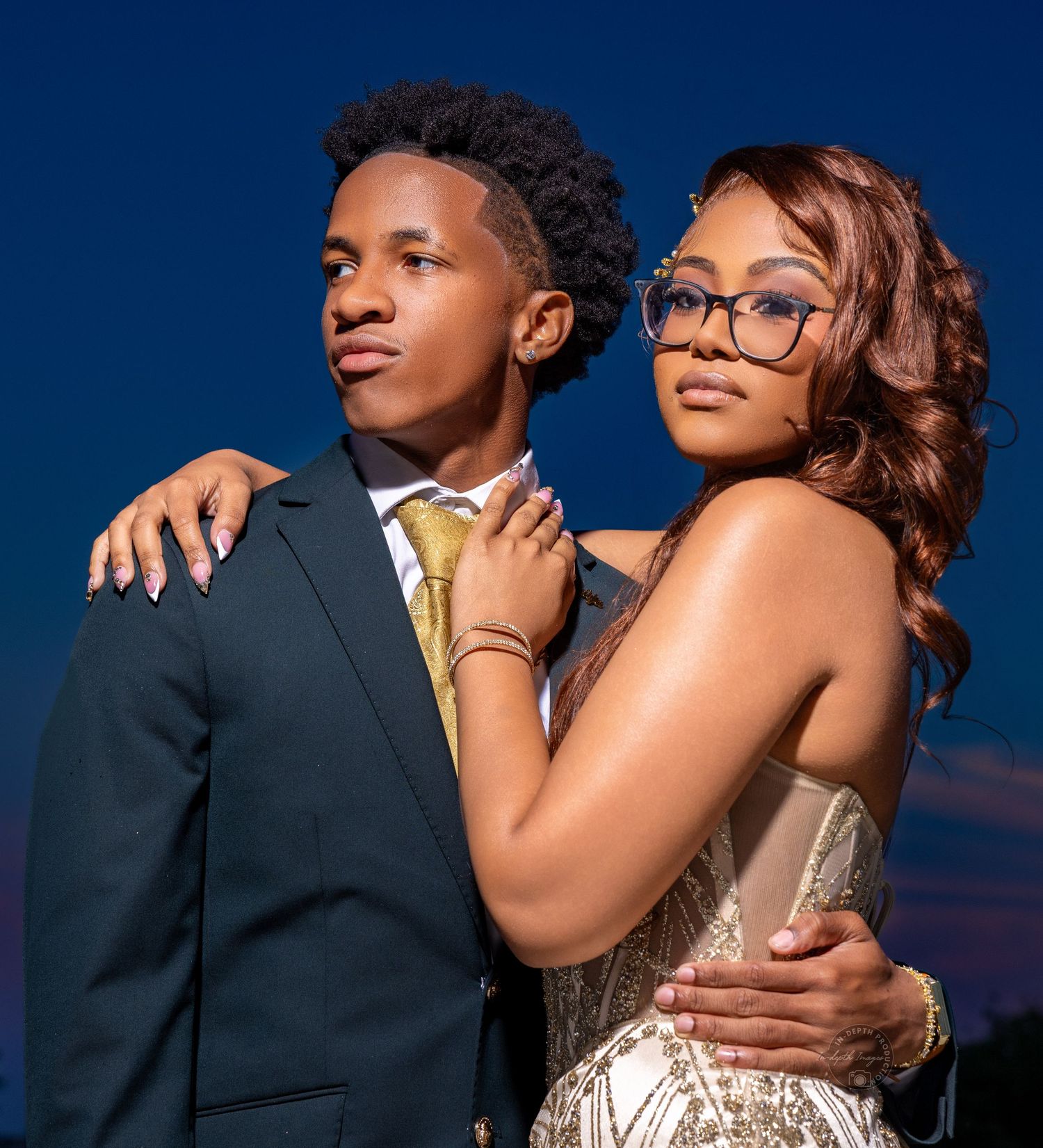 A young couple dressed in formal attire poses elegantly against a twilight blue sky for prom photos.