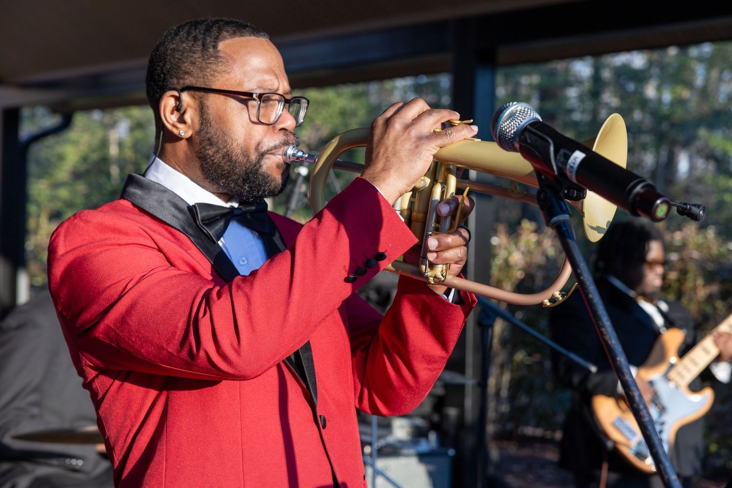 Man in red blazer playing trumpet on stage with band performing live music outdoors.