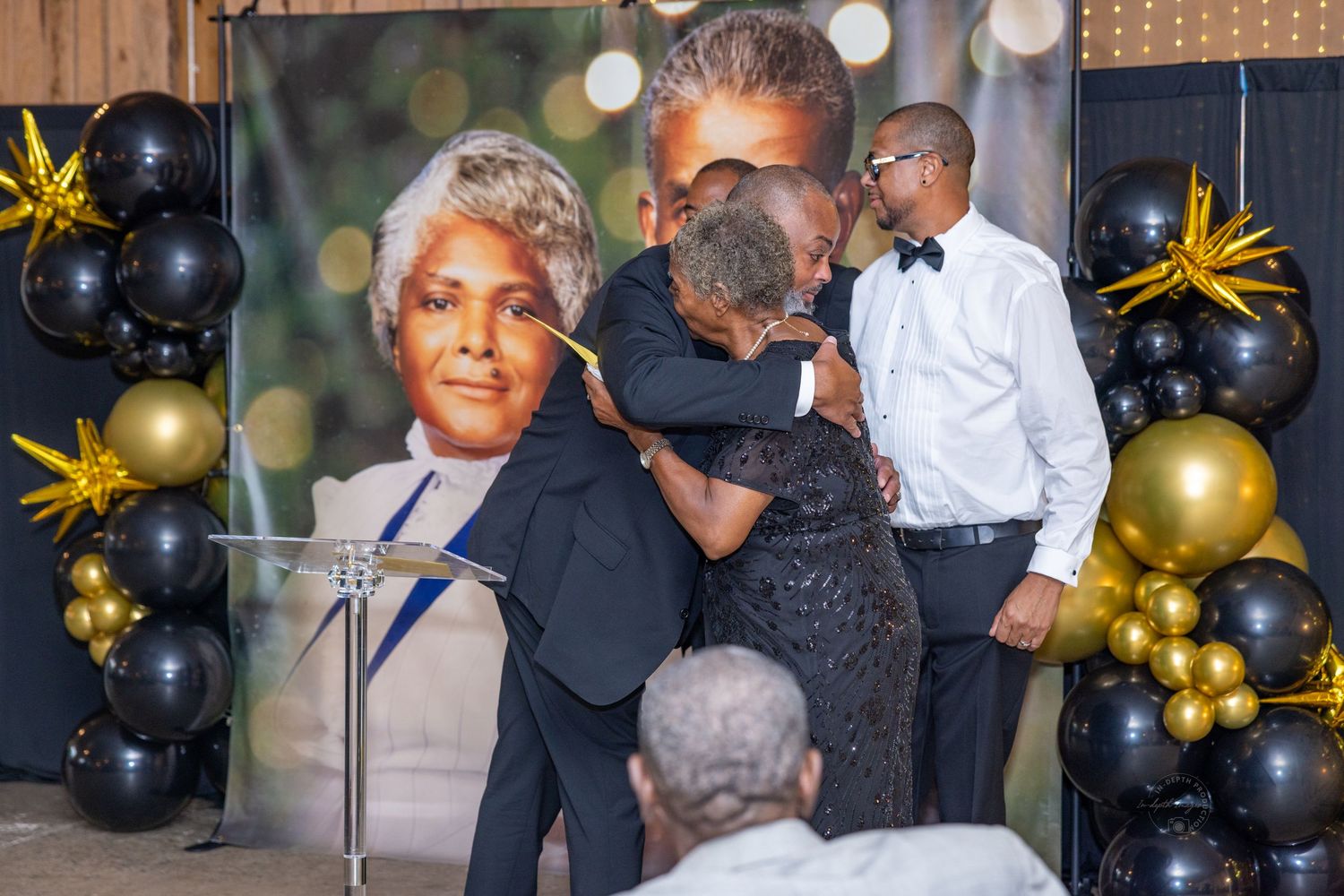 People hugging at a memorial celebration with black and gold balloons and a portrait of an elderly woman.