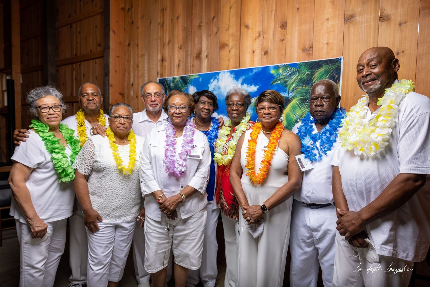 A diverse group of people wearing white outfits and colorful floral leis pose together for a group photo indoors.