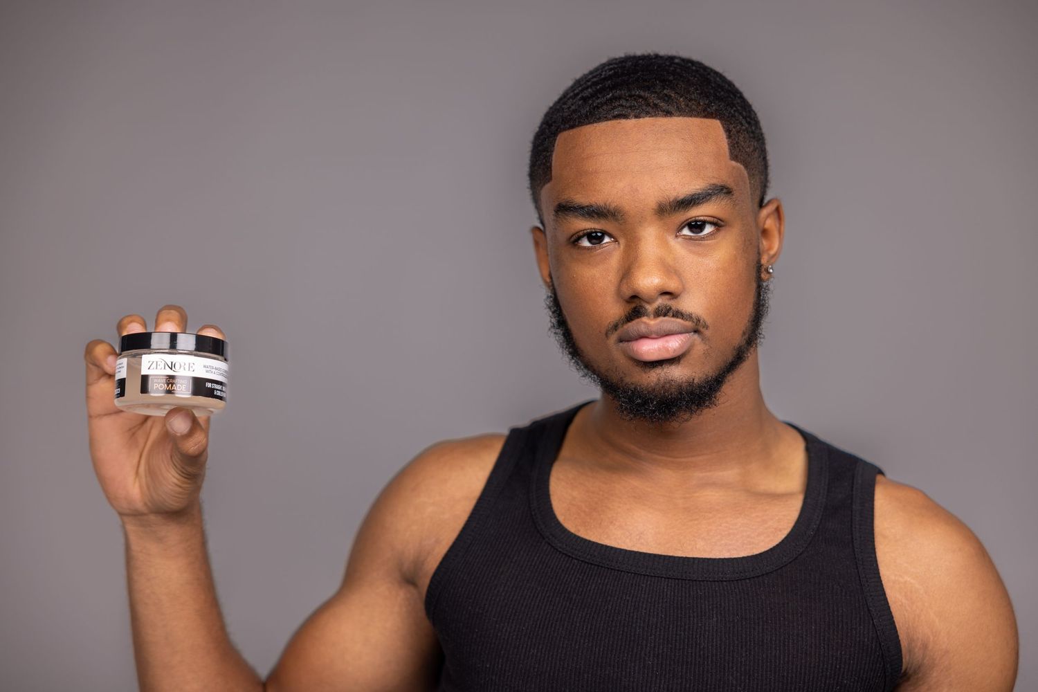 A young Black man in a black tank top holds a small product container against a gray studio background.