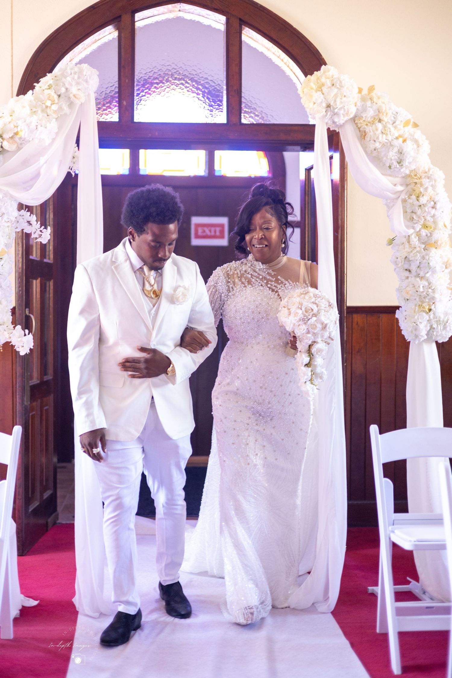 Bride and groom walk down the aisle in white wedding attire under a floral arch in a church.