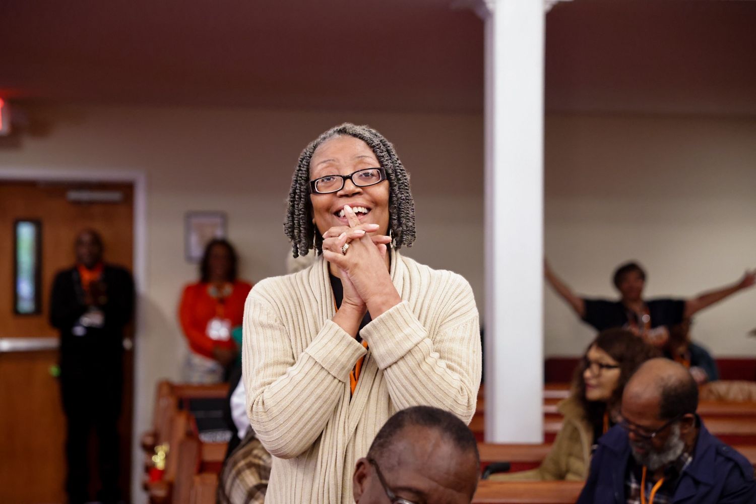 Smiling woman with gray locs and glasses clasps her hands in prayer during a church service while worshippers raise their arms behind her.