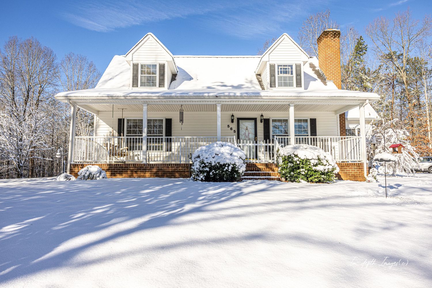 A beautiful white farmhouse with wrap-around porch covered in snow under a clear blue winter sky.