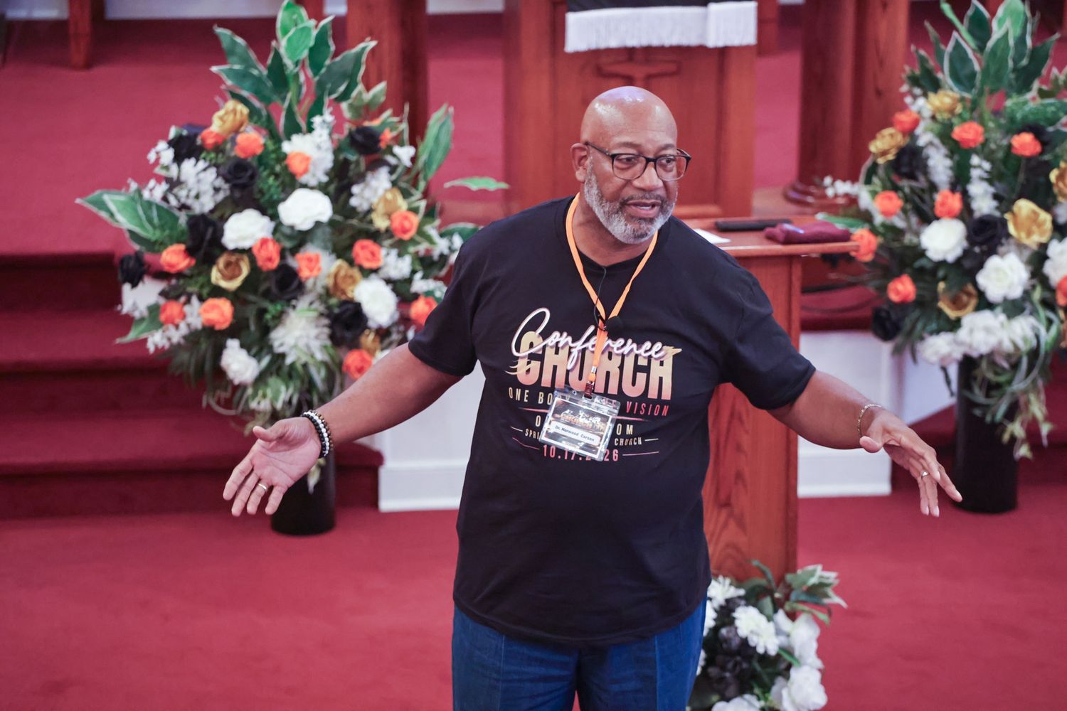 Bearded man in glasses and a black Church t-shirt gestures with open arms near floral arrangements in a red-carpeted sanctuary.