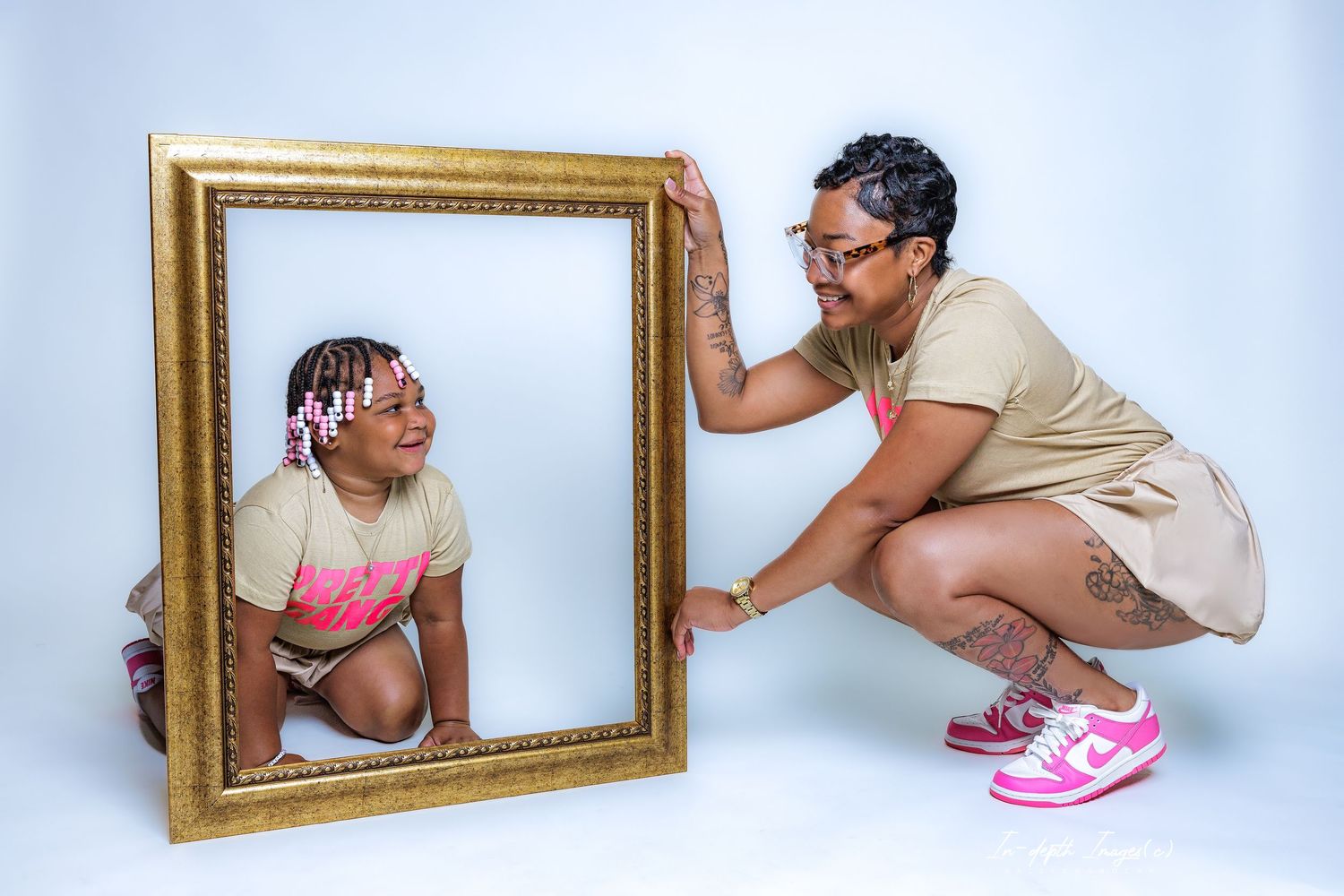Woman crouching beside ornate gold frame while young girl poses inside during creative studio photo session.