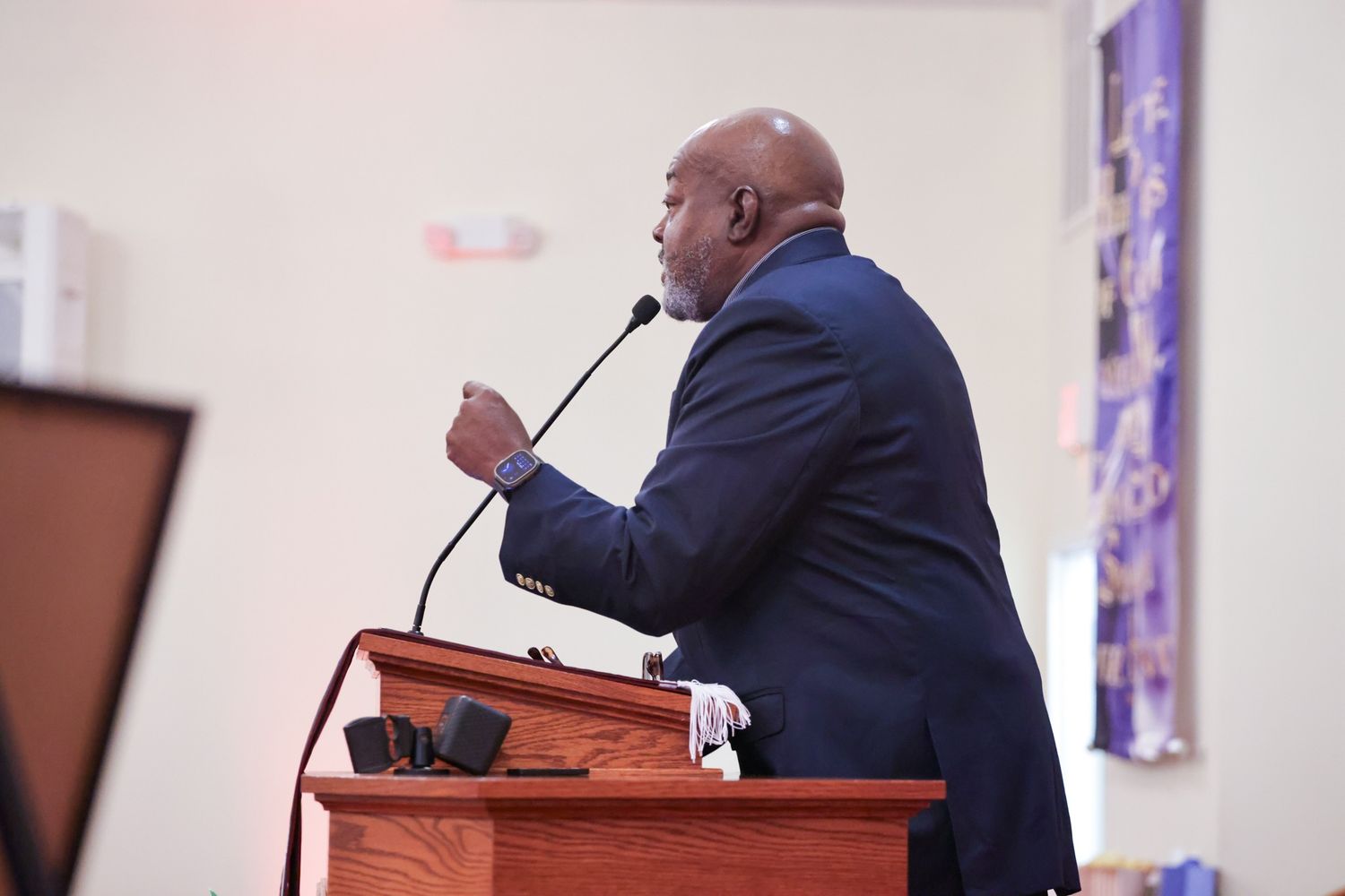 Man in a dark suit speaks into a microphone while gesturing from a wooden pulpit during a church service.