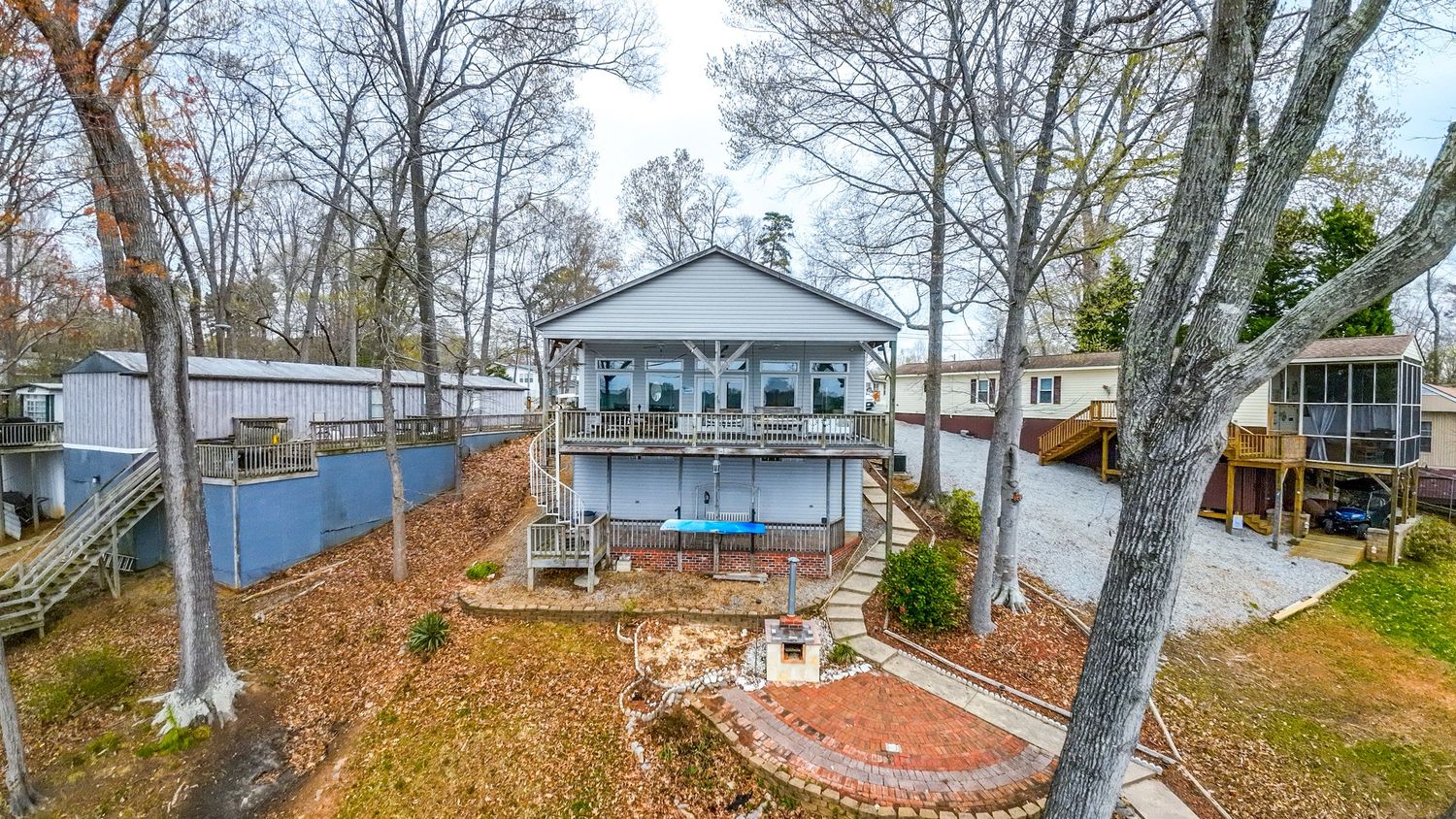 Cozy waterfront cabin featuring a spacious deck, fire pit area, and neighboring properties visible through bare winter trees.
