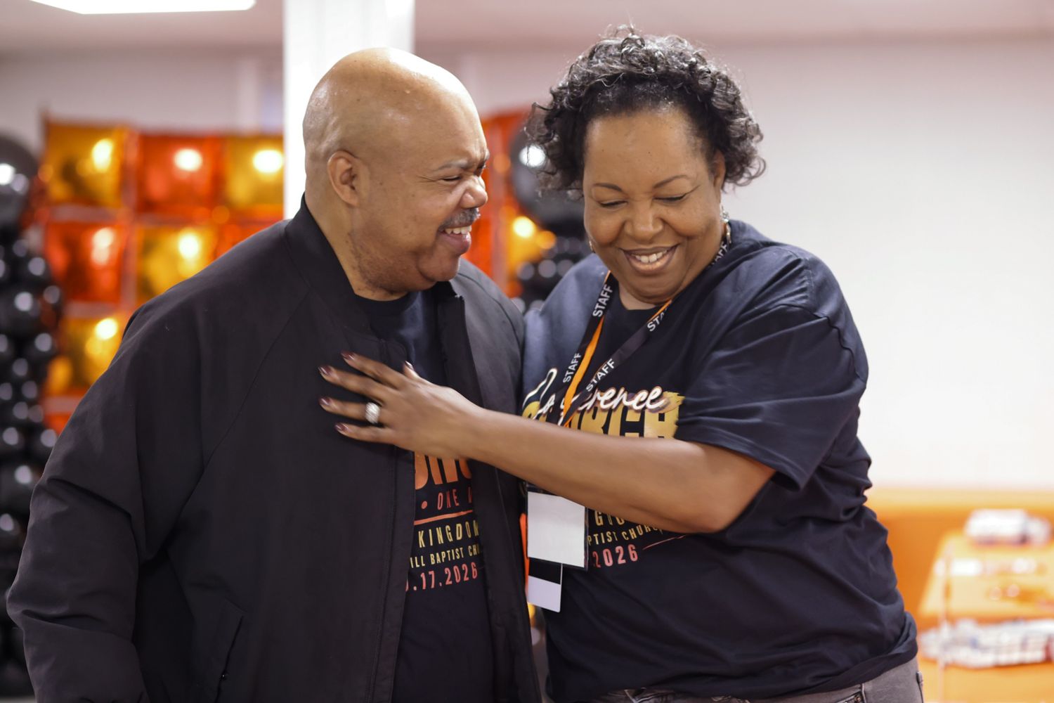 Two smiling people in dark graphic t-shirts share a warm, joyful moment, with one resting a hand on the other's chest at an indoor event.