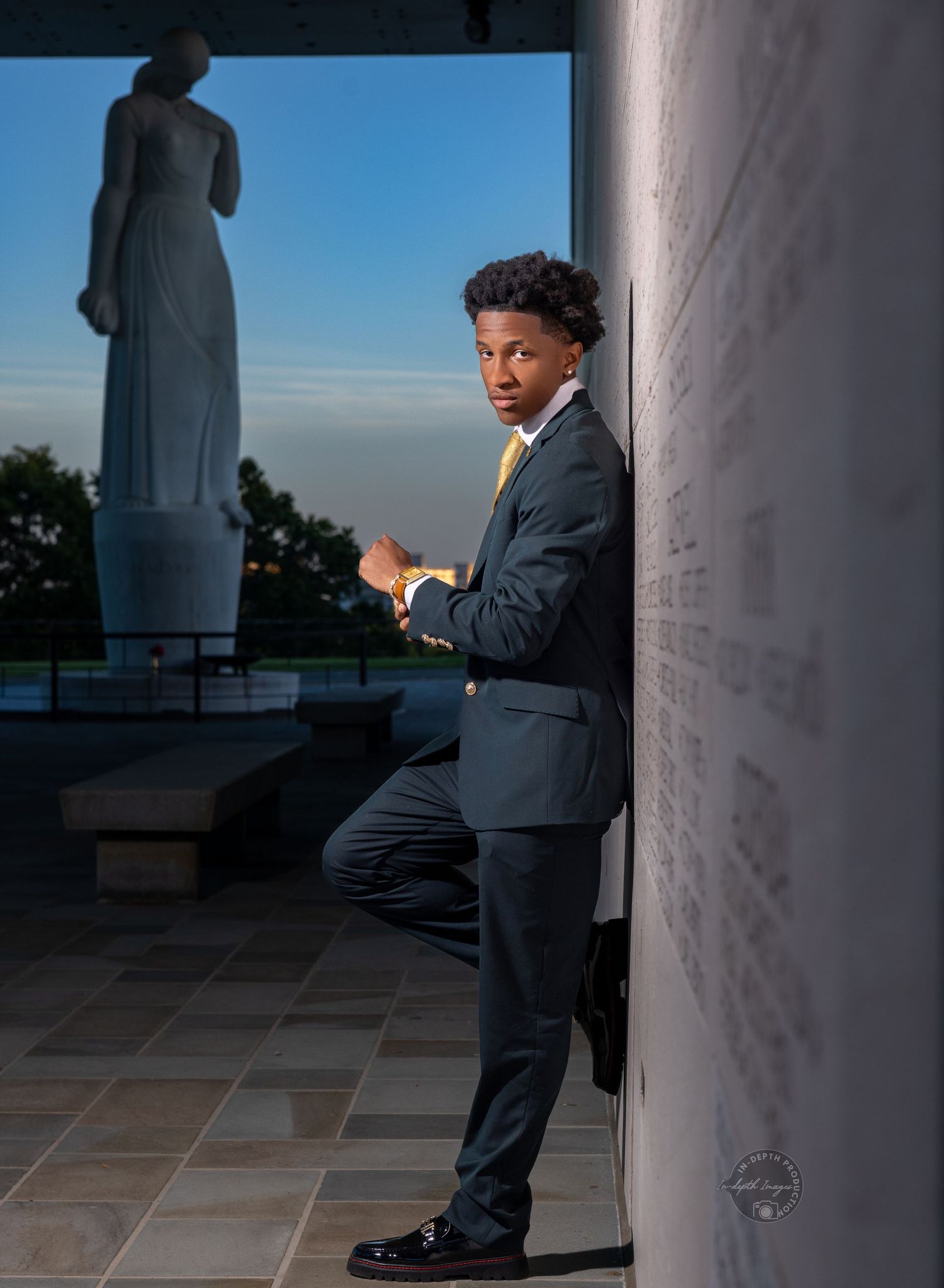 Young man in gray suit with yellow tie poses confidently against a dark wall near a large statue at dusk.