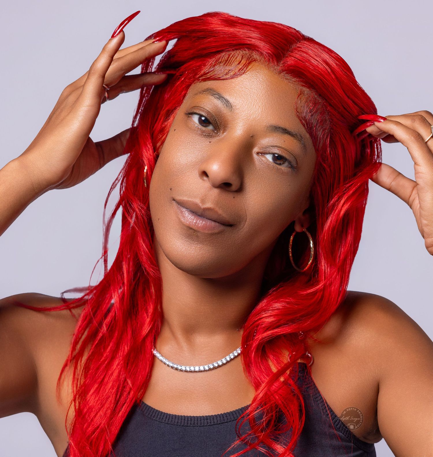 Woman with vibrant red long wavy hair, red nails, gold hoop earrings, and pearl necklace posing confidently.