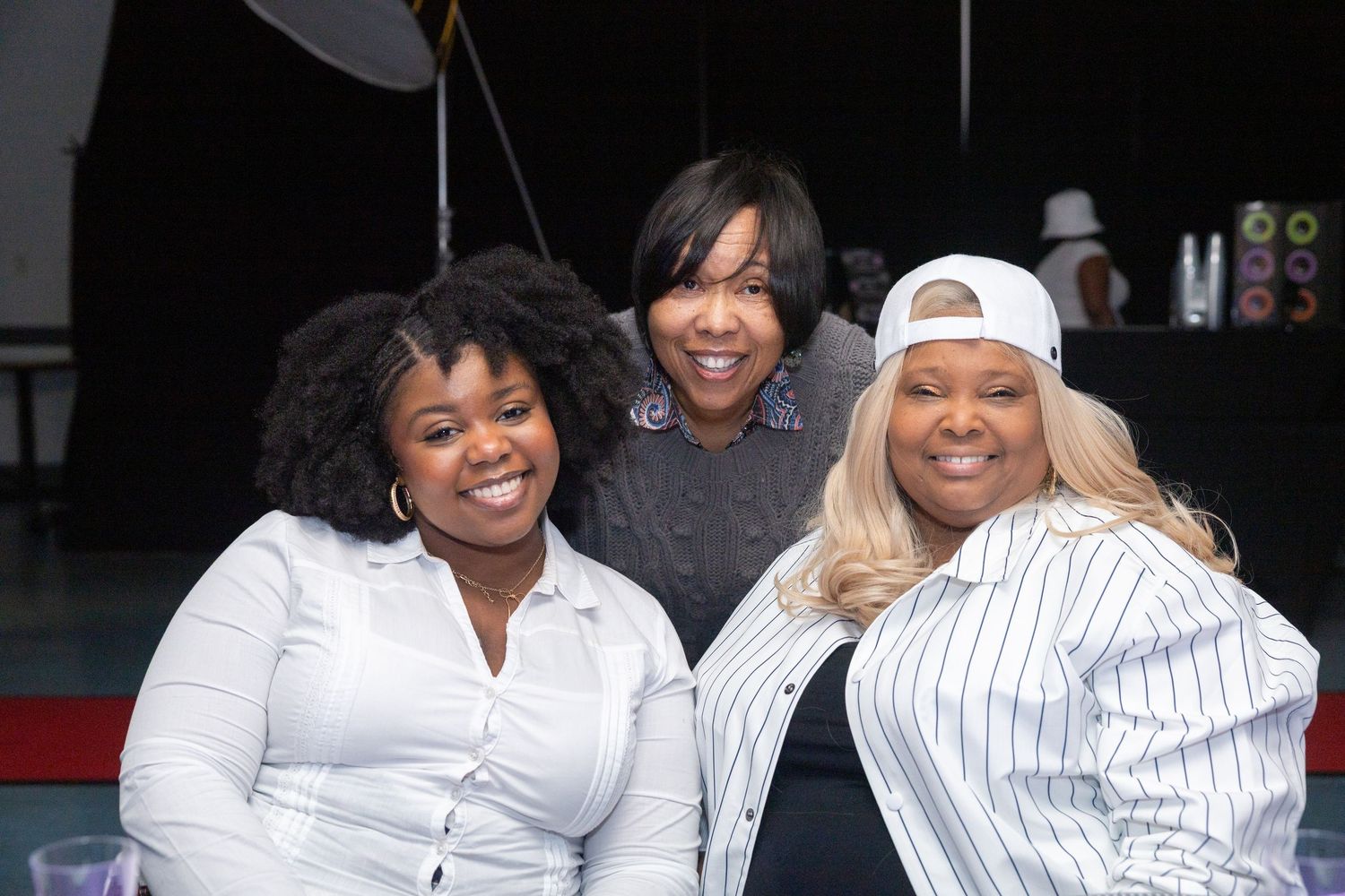 Three women smiling together at an indoor event, posing for a group photo in a warmly lit venue.