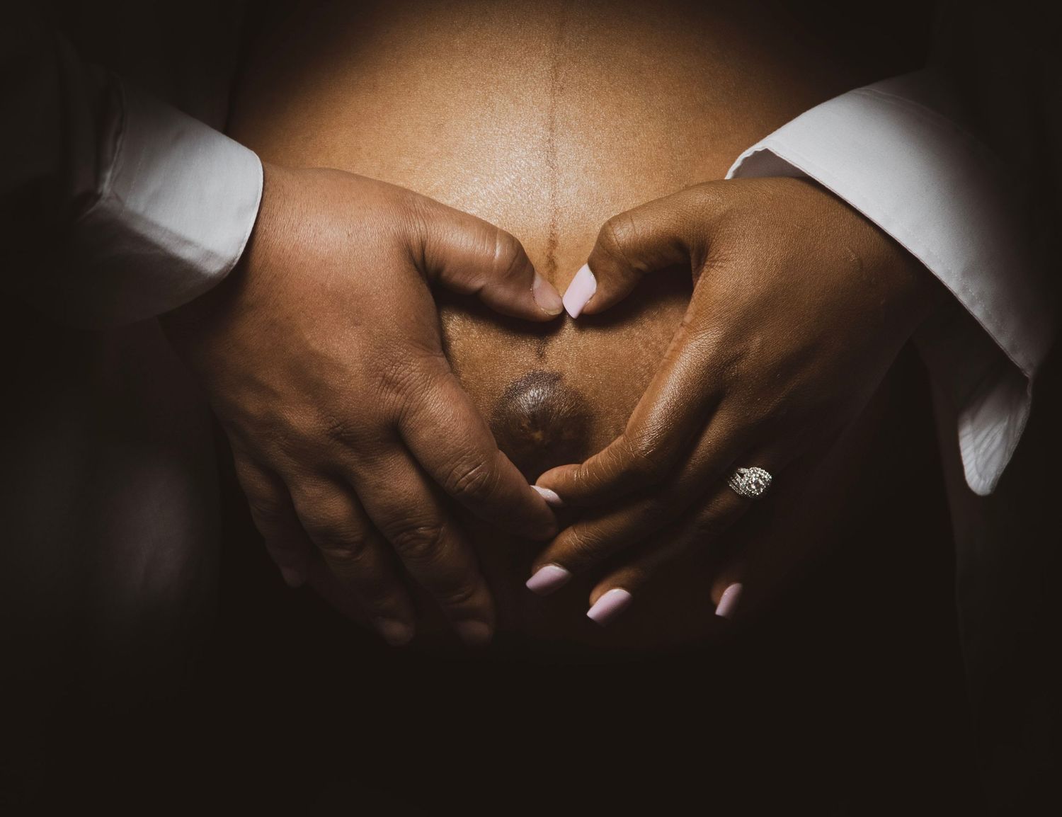 Two hands forming a heart shape over a pregnant belly with engagement ring visible.